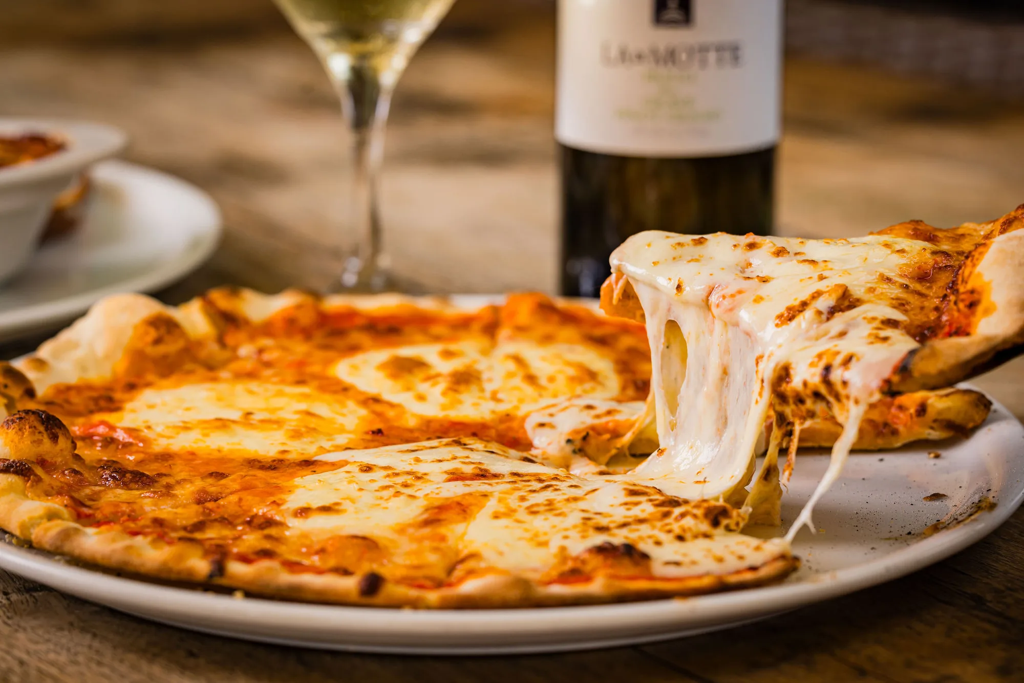 A slice being lifted from a cheese pizza, with a glass of white wine and a bottle behind it