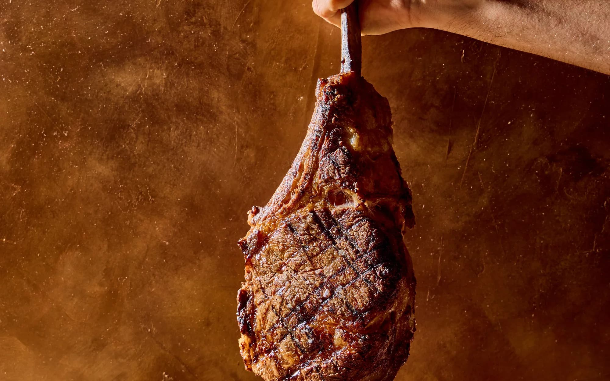 A hand holding up a charcoal-grilled tomahawk steak by its bone against a copper background
